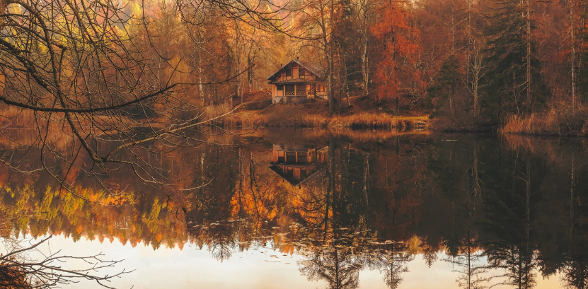 A scene of a cabin centered in a forest with fall foliage. A reflection of the scene is mirrored from a still lake.