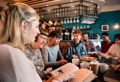 a woman on the left holds a book. people around a table are having a discussion